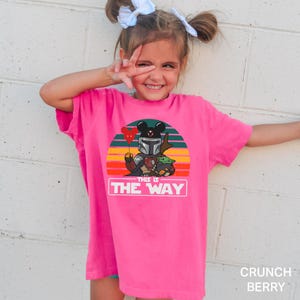 May include: A bright pink t-shirt featuring a graphic of a Mandalorian-inspired character with Mickey Mouse ears, Grogu, and the text "THIS IS THE WAY" against a sunset background. The shirt is labeled "Crunch Berry".