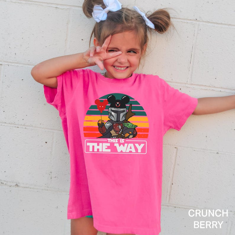 May include: A bright pink t-shirt featuring a graphic of a Mandalorian-inspired character with Mickey Mouse ears, Grogu, and the text "THIS IS THE WAY" against a sunset background. The shirt is labeled "Crunch Berry".