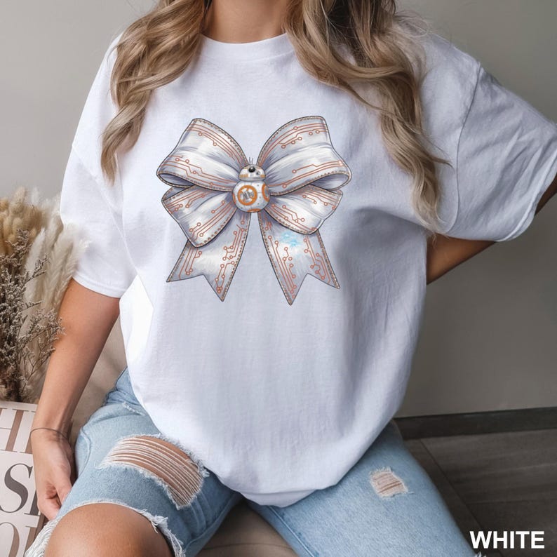 Puede incluir: Camiseta blanca con un dise&ntilde;o de lazo grande con patrones de placa de circuito y un personaje BB-8 en el centro. El lazo tiene una combinaci&oacute;n de colores azul claro y cobre. La palabra "WHITE" es visible en la esquina inferior derecha.