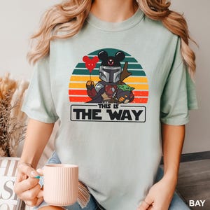 May include: A light green t-shirt featuring a cartoon design of a Mandalorian character with a Mickey Mouse head, holding a red heart-shaped balloon, and Grogu. The design is set against a sunset background with the text "THIS IS THE WAY".
