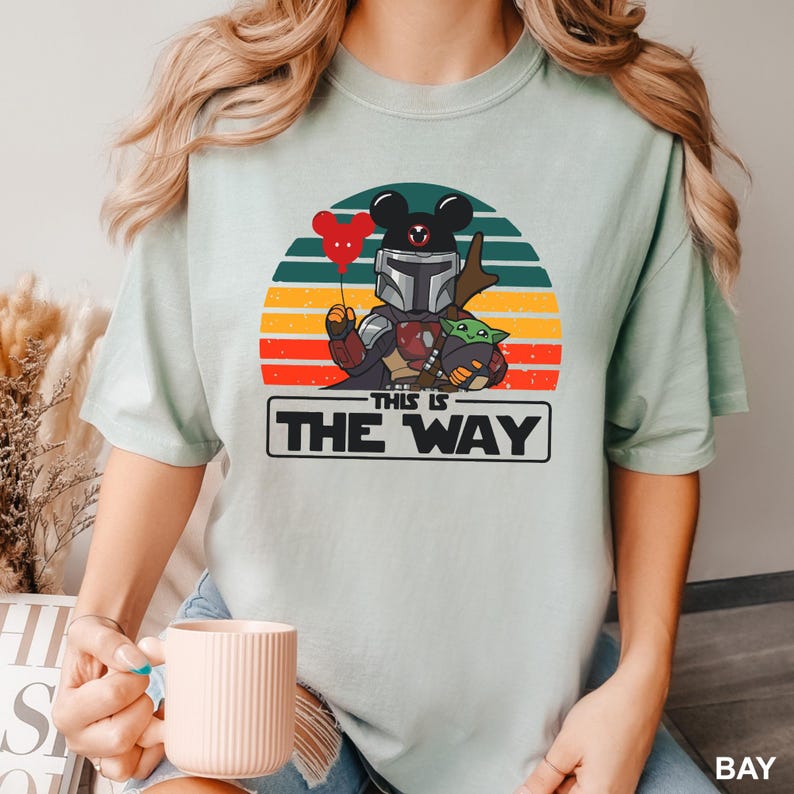 May include: A light green t-shirt featuring a cartoon design of a Mandalorian character with a Mickey Mouse head, holding a red heart-shaped balloon, and Grogu. The design is set against a sunset background with the text "THIS IS THE WAY".