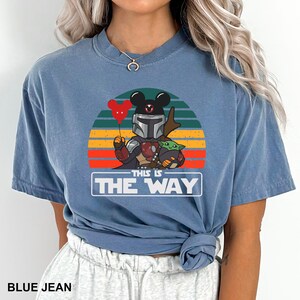 May include: Blue jean-colored t-shirt featuring a graphic of a Mandalorian-inspired character with Mickey Mouse ears, holding a red heart balloon, and Grogu. The text "THIS IS THE WAY" is displayed below the graphic.