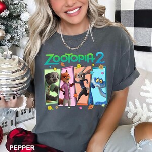 May include: Dark gray t-shirt featuring the text "ZOOTOPIA 2" in teal and green. Below the text are four rectangular panels with cartoon characters from the movie. The shirt has rolled-up sleeves.