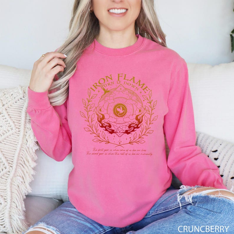May include: A long-sleeved, pink sweatshirt with a gold graphic design. The design includes the text "IRON FLAME TURN IT DOWN" around a circular emblem with a bird and flames. Additional text is below the emblem.