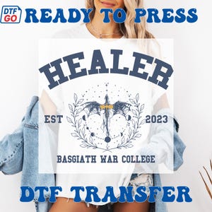May include: White t-shirt featuring the word "HEALER" in navy blue arched text. The design includes a dragon illustration with celestial elements. The text "EST 2023" and "BASGIATH WAR COLLEGE" are also visible on the shirt.