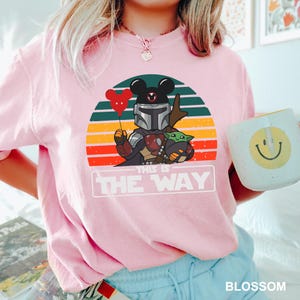 May include: Pink t-shirt featuring a graphic of The Mandalorian with a Mickey Mouse head, holding a red heart balloon, and Grogu. The design is set against a retro sunset with the text "THIS IS THE WAY". A white mug with a yellow smiley face is also visible.