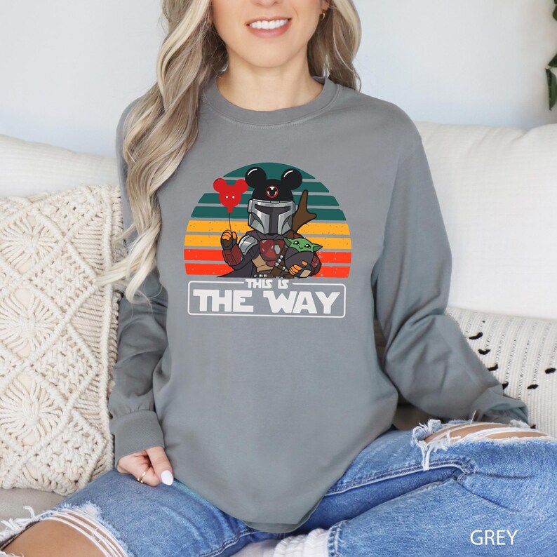 May include: A grey long-sleeve shirt featuring a graphic of a Mandalorian-Mickey Mouse hybrid holding a red heart balloon and Grogu. The text "THIS IS THE WAY" is below the graphic. The shirt is a comfortable, casual style.