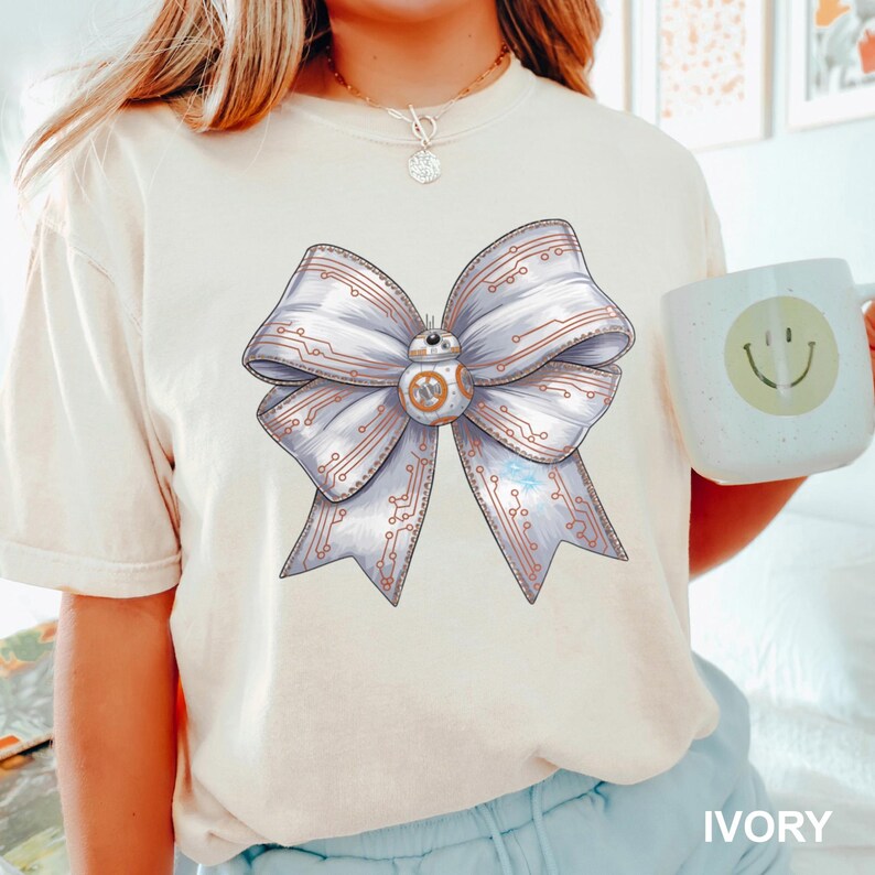 Puede incluir: Camiseta color marfil con un dise&ntilde;o de lazo grande y detallado. El lazo es blanco con detalles de placa de circuito y un droide BB-8 en el centro. La palabra "IVORY" est&aacute; impresa en la parte inferior.