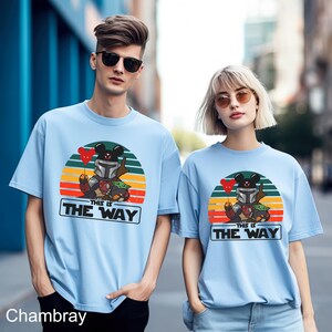 May include: Light blue t-shirts featuring a cartoon design of a character in a helmet holding a red heart balloon, with a green creature and the text "THIS IS THE WAY" in a white rectangle. The design is set against a sunset background.