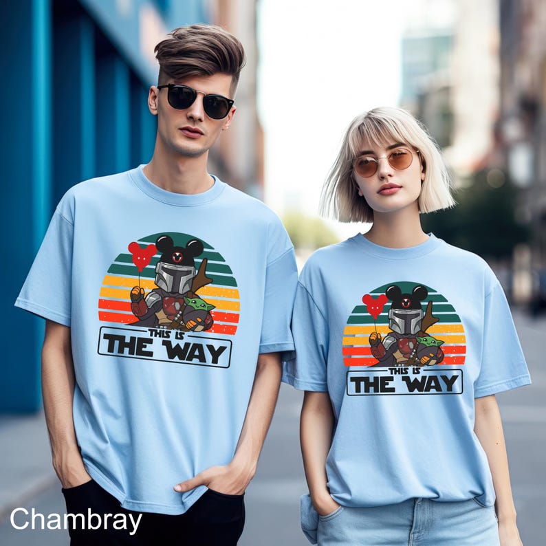 May include: Light blue t-shirts featuring a cartoon design of a character in a helmet holding a red heart balloon, with a green creature and the text "THIS IS THE WAY" in a white rectangle. The design is set against a sunset background.