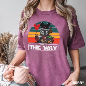 May include: Berry-colored t-shirt featuring a graphic of a Mandalorian-inspired character with a Mickey Mouse-shaped helmet and a Grogu figure. The design includes a red balloon and the text "THIS IS THE WAY" in a white rectangle.