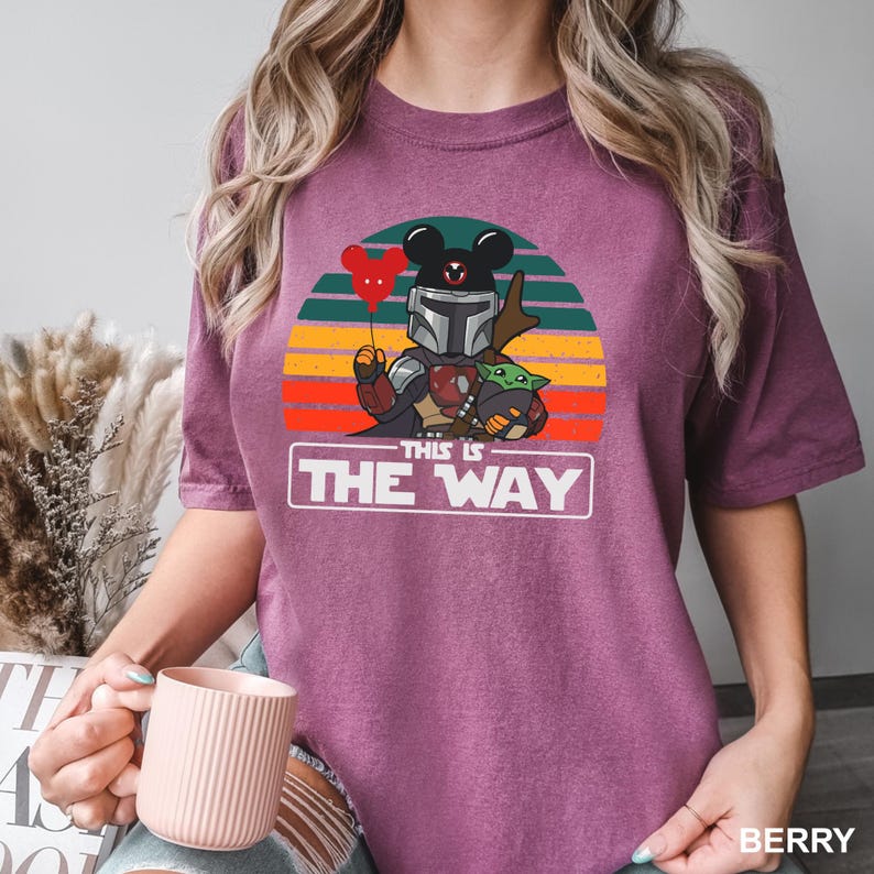May include: Berry-colored t-shirt featuring a graphic of a Mandalorian-inspired character with a Mickey Mouse-shaped helmet and a Grogu figure. The design includes a red balloon and the text "THIS IS THE WAY" in a white rectangle.