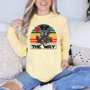 May include: Butter yellow long-sleeve shirt featuring a graphic of a Mandalorian-inspired character with Mickey Mouse ears holding a heart balloon, and Grogu. The text "THIS IS THE WAY" is displayed below the graphic.