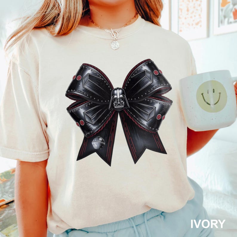 Puede incluir: Camiseta de color marfil con un dise&ntilde;o de lazo grande y detallado. El lazo es negro con detalles rojos y una imagen central del casco de Darth Vader. La camiseta tiene mangas cortas y cuello redondo. La palabra "IVORY" est&aacute; impresa en la parte inferior.