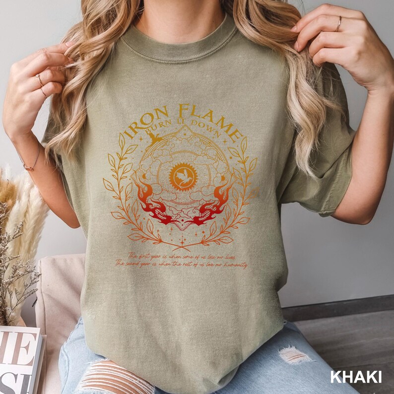 May include: Khaki-colored t-shirt with a circular graphic featuring the text "IRON FLAME TURN IT DOWN." The design includes a central emblem, flames, and botanical elements in orange and red. Below the graphic are two lines of text.