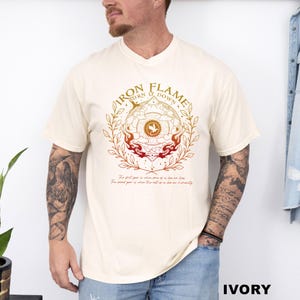 May include: Ivory-colored t-shirt featuring a circular design with the words "IRON FLAME TURN IT DOWN" in a gold font. The design includes a central emblem with a bird and flames, and text at the bottom. The shirt is a relaxed fit.