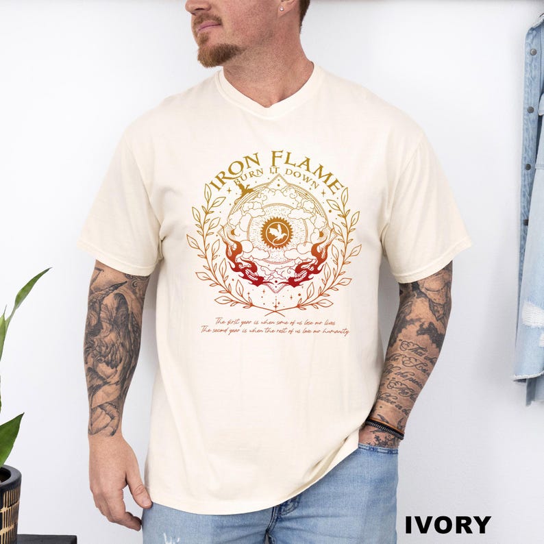 May include: Ivory-colored t-shirt featuring a circular design with the words "IRON FLAME TURN IT DOWN" in a gold font. The design includes a central emblem with a bird and flames, and text at the bottom. The shirt is a relaxed fit.