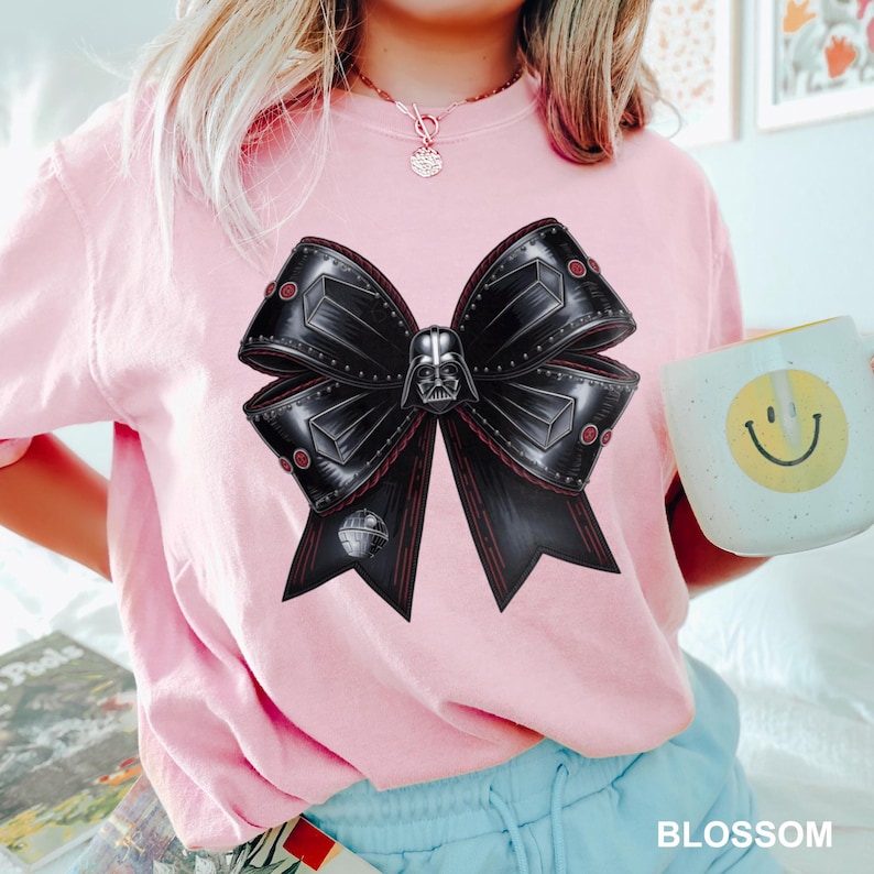 Puede incluir: Camiseta rosa claro con un gran dise&ntilde;o de lazo negro con un gr&aacute;fico de Darth Vader. El lazo tiene detalles rojos y un gr&aacute;fico de la Estrella de la Muerte. La camiseta es de color rosa claro.