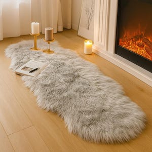 Puede incluir: Una larga alfombra de piel sintética gris se encuentra sobre un suelo de madera, cerca de una chimenea. Dos velas están sobre soportes de madera, y un libro con un bolígrafo descansa sobre la alfombra. También se ve una vela encendida.