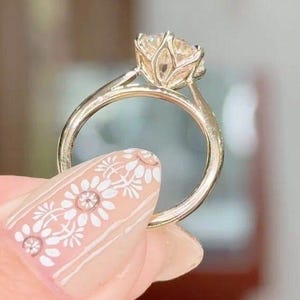 May include: A gold ring with a large, clear gemstone. The ring's setting has a floral design, and the band is smooth. The ring is held up by a hand with white floral nail art.
