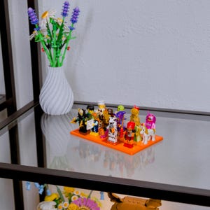 May include: A collection of colorful Lego minifigures displayed on an orange base. The figures include various characters with unique designs. A white vase with a spiral design holds artificial flowers with purple and white blooms.