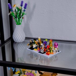 May include: A display of Lego minifigures on a white base, featuring various characters with different outfits and accessories. A white vase with a spiral design holds a bouquet of artificial flowers. The display sits on a glass shelf.