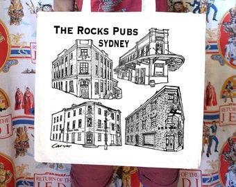 May include: A white tote bag featuring black line drawings of four pubs in Sydney, Australia. The text "The Rocks Pubs Sydney" is printed in black at the top of the bag.