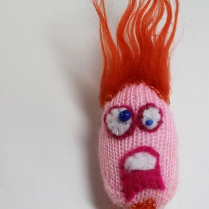 May include: A pink knitted character with a red yarn wig and a pink and white mouth. The character has blue eyes and a white background.