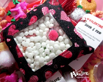 May include: A black and pink fabric pouch with a clear window, filled with white styrofoam packing peanuts. The pouch has a heart pattern and the text "VALENTINE Seek & Find" is printed on a white tag attached to the pouch.