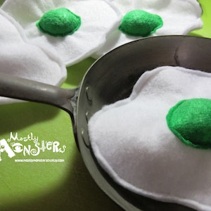 May include: A set of felt toy eggs with green yolks in a black frying pan. The eggs are white and have a realistic appearance. The pan is empty except for the eggs.