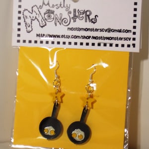 Puede incluir: Un par de pendientes colgantes con una sartén negra en miniatura con dos huevos amarillos en su interior. La sartén está unida a un gancho plateado con un amuleto de estrella amarilla.