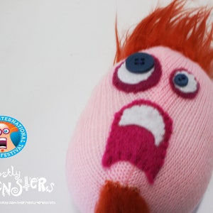 May include: A pink knitted monster doll with orange hair and a surprised expression. The doll has button eyes, a magenta mouth, and a white open mouth. The San Diego International Fringe Festival logo is visible.