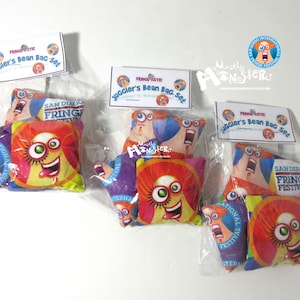 May include: Three sets of bean bags in clear plastic bags. Each set contains two bean bags with colorful designs featuring a cartoon character with a large eye and a wide mouth. The bean bags are labeled "Juggler's Bean Bag Set" and "San Diego Fringe Festival".