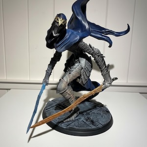 May include: A detailed figurine of a warrior in dark armour, a blue cape, and a mask. The figure is holding two swords, one blue and one brown, and is posed on a circular base. The warrior's helmet has gold accents.