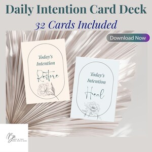 May include: A card deck titled "Daily Intention Card Deck" with the text "32 Cards Included." Two cards are visible, one reading "Restore" and the other "Heal." Each card features floral illustrations and the words "Today's Intention."