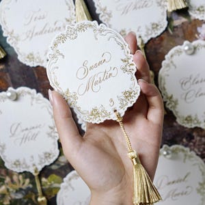 May include: Elegant white place cards with gold calligraphy and decorative floral borders. Each card features a gold tassel and pearl accent. The name "Susan Martin" is visible on the front of the card. Perfect for weddings or special events.