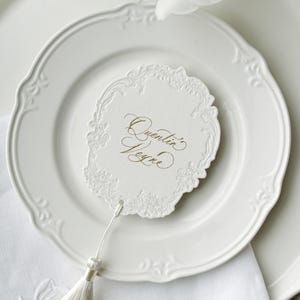 Peut inclure: Une présentation de table blanche avec une assiette décorative, une serviette blanche et un carton de table rond. Le carton de table porte l'inscription dorée "Quentin Peyre" et un pompon blanc. L'assiette a un bord festonné.