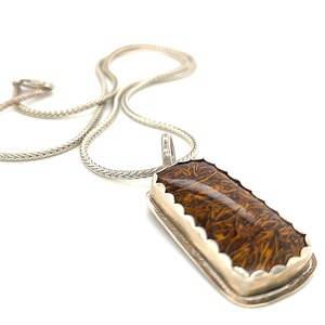 May include: A silver chain necklace with a brown and gold rectangular pendant. The pendant has a scalloped edge and a swirling pattern.