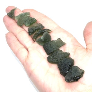 May include: A collection of green, rough, and irregularly shaped Moldavite crystals. The crystals are arranged in a line on a hand.