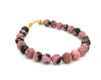 Puede incluir: Un brazalete de piedras preciosas rosa y negro con un cierre de oro. El brazalete está hecho de cuentas redondas.