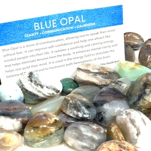 May include: A collection of blue opal tumbled stones. The stones are a mix of gray, white, and brown with black veins. The stones are smooth and polished. The image includes a card with the text "Blue Opal Clarity Communication Calmness".