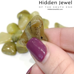 May include: A close-up of a light green and brown agate stone with brown and white banding. The stone is being held by a hand with purple nail polish.
