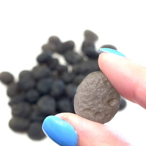 May include: A close-up of a gray, smooth, oval-shaped sea glass stone with a textured surface. The stone is being held between two fingers.