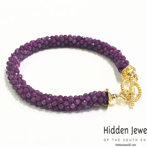 Op de afbeelding: Een armband van paarse edelstenen met een gouden sluiting. De armband is gemaakt van kleine, ronde kralen en heeft een eenvoudig, elegant ontwerp.