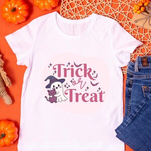 Puede incluir: Una camiseta blanca de manga corta con un estampado gráfico de un fantasma con un sombrero de bruja morado que sostiene un pequeño gato morado. El texto "Trick or Treat" está impreso en morado, junto a pequeñas ilustraciones de estrellas y murciélagos.