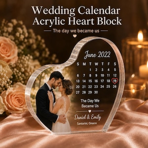 May include: A heart-shaped clear acrylic block featuring a printed photograph of a couple, a June 2022 calendar with the 19th circled in red, the text The Day We Became Us, the names Daniel & Emily, and the location Santorini, Greece.