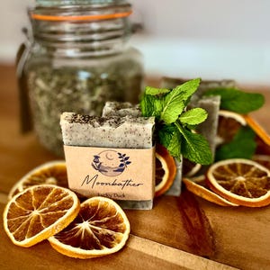 May include: Handmade soap bars with a kraft paper label that reads "Moonbather Lucky Duck". The soap is light green with dark flecks, and is accompanied by dried orange slices, fresh mint sprigs, and a jar of green herbs.