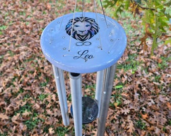 May include: A light blue wind chime with a Leo zodiac sign design. The top disc features a lion's head illustration, stars, and the word "Leo". The chime has silver-colored tubes and a black circular clapper, hanging outdoors among fallen leaves.