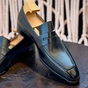 May include: Black leather penny loafers with a glossy finish. The shoes have a classic design with a square toe and a strap across the vamp. A wooden shoe tree is inserted inside the shoe. The loafers are on a dark surface.