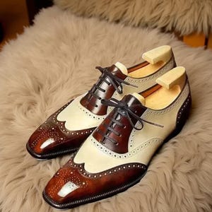 May include: A pair of two-tone leather dress shoes with brown and cream-colored panels. The shoes feature decorative perforations and dark brown laces. Shoe trees are inserted in the shoes.